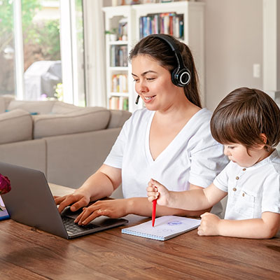Woman working from home with headset on laptop with her son coloring next to her.